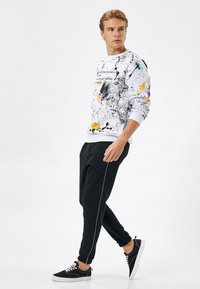 Vit sweatshirt med svart, gul och teal färgstänkdesign, kombinerat med svarta joggers med en vit sidostrip och svarta sneakers.