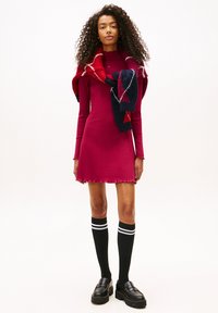 Κόκκινο φορεμά ριπ με μήκος μέχρι το γόνατο και κυματιστό τελείωμα. Συνδυασμένο με κόκκινο και ναυτικό κασκόλ και μαύρες καλτσες έως το γόνατο με λευκές ρίγες. Μαύρα loafers.