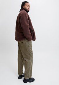 Bruine fleec Jas met een ritszak, gecombineerd met khaki corduroy broeken en zwarte schoenen, getoond op een mannelijke model vanuit een zijaanzicht.