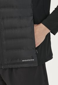 Schwarze gepolsterte Weste mit glatter Oberfläche, die eine Reißverschlusstasche und das "ENDURANCE"-Logo am unteren Saum aufweist. Das Material ist leicht und strapazierfähig.