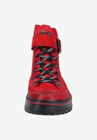 Botte montante rouge avec des lacets noirs et blancs, une large sangle avec une boucle métallique, et une semelle noire épaisse avec crampons.