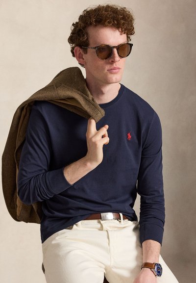 Egy férfi barna napszemüveget visel, tengerészkék hosszú ujjú inget piros logóval, bézs nadrágot, barna övet, barna dzsekit tart a vállán.