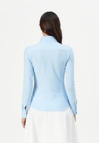 Camicia a maniche lunghe a righe azzurre con colletto a punta e orlo curvo. Presenta polsini con chiusura a bottone. Texture del tessuto liscia.
