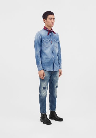 Man wearing faded blue denim shirt, distressed jeans, red neck scarf, and black leather ankle boots standing against white background.