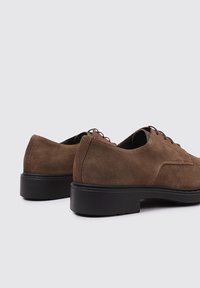 Zapatos de ante marrón con punta redonda, suela baja negra y un diseño de cordones minimalista. Presentan una textura suave y líneas limpias.
