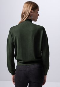 Suéter de color verde oscuro con cuello alto, corte holgado, puños acanalados y una textura suave. Combinado con jeans oscuros que tienen dos bolsillos.