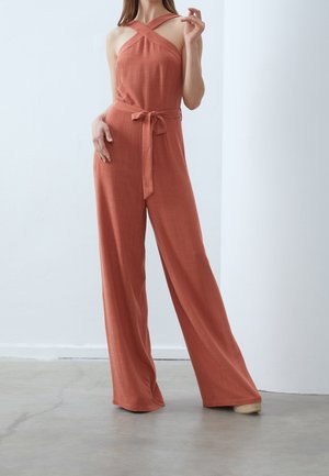 Kvinde iført rustfarvet, ærmeløs buksedragt med krydsede stropper og bundet taljebælte, stående indendørs på betongulv.