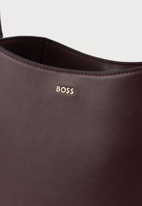 Bordó bőr kézitáska sima felülettel, arany "BOSS" logóval és nyitott felső kialakítással.