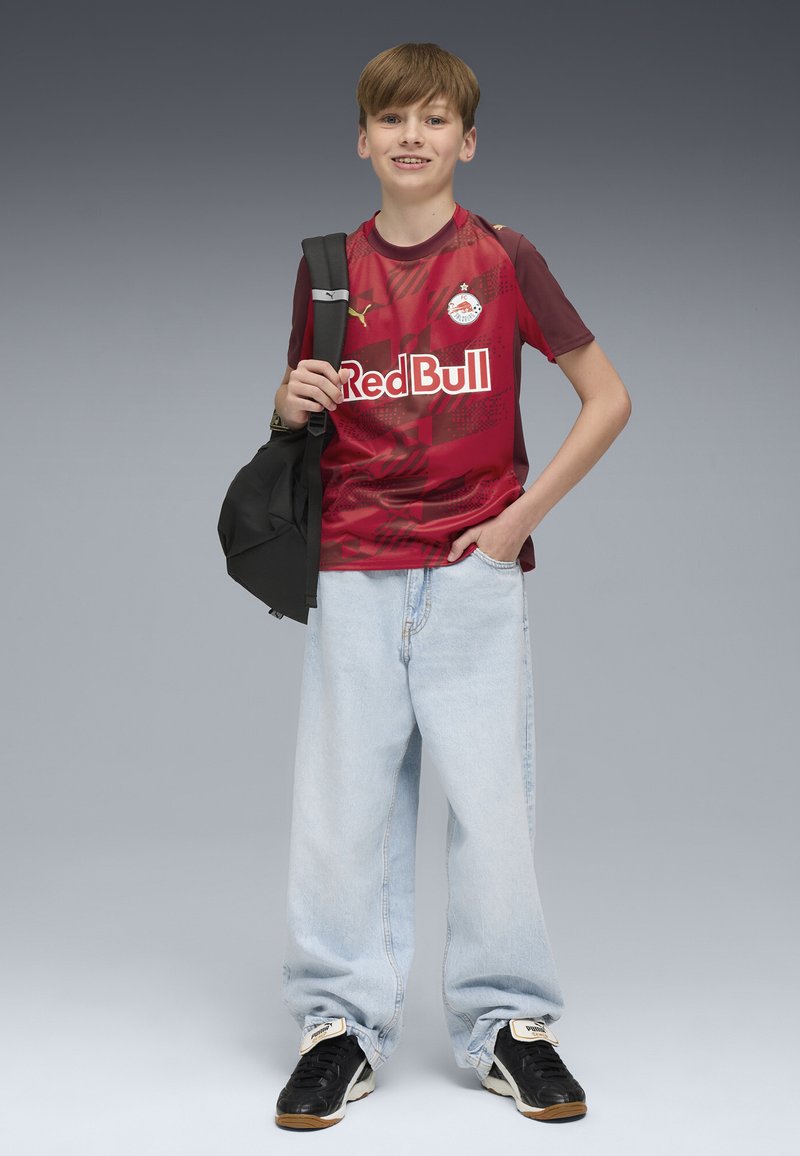 Camiseta de fútbol roja de manga corta, con un degradado de burdeos a rojo, que presenta el logotipo de "Red Bull"; combinada con unos vaqueros anchos de color azul claro y zapatillas negras.