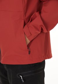 Röd vattentät jacka med en slät textur, en dragkedja fram och justerbara ärmslut. Har sidofickor och en dragsko i fållen.
