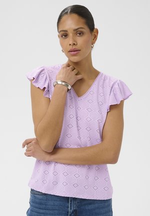 Femme aux cheveux foncés attachés en chignon portant un haut lilas à motifs avec des manches à volants, un jean bleu, un bracelet argenté et des petites boucles d'oreilles créoles.