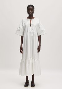 Witte katoenen jurk met korte puff mouwen, een V-hals met strikdetail en een gelaagde kraag met ruffles. Model draagt zwarte schoenen.