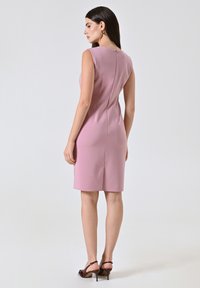 Figurbetontes pinkfarbenes ärmelloses Kleid mit einem Reißverschluss auf der Rückseite und einem Schlitz. Glatter Stoff mit klarer Silhouette, knielang, kombiniert mit Riemchenschuhen.