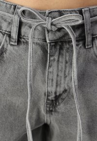 Pantalones de mezclilla gris con cintura ajustable mediante cordón. Presentan un aspecto texturizado y ligeramente desvanecido, con un lazo atado en la parte frontal.