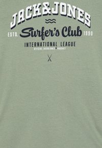Svijetlozelena pamučna majica s podebljanim crnim tekstom. Dizajn uključuje "JACK & JONES", "Surfer's Club" i ukrštene daske za surfanje, s mekim teksturom.