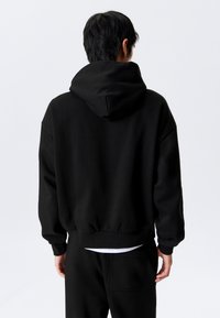 Schwarzer Kapuzenpullover mit lockerer Passform, gerippten Ärmelbündchen und Saum. Glatte Textur, minimalistisches Design, keine sichtbaren Muster oder Akzente.