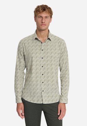 Man draagt een overhemd met lange mouwen en een licht geometrisch patroon en een donkere broek, staand tegen een effen witte achtergrond.