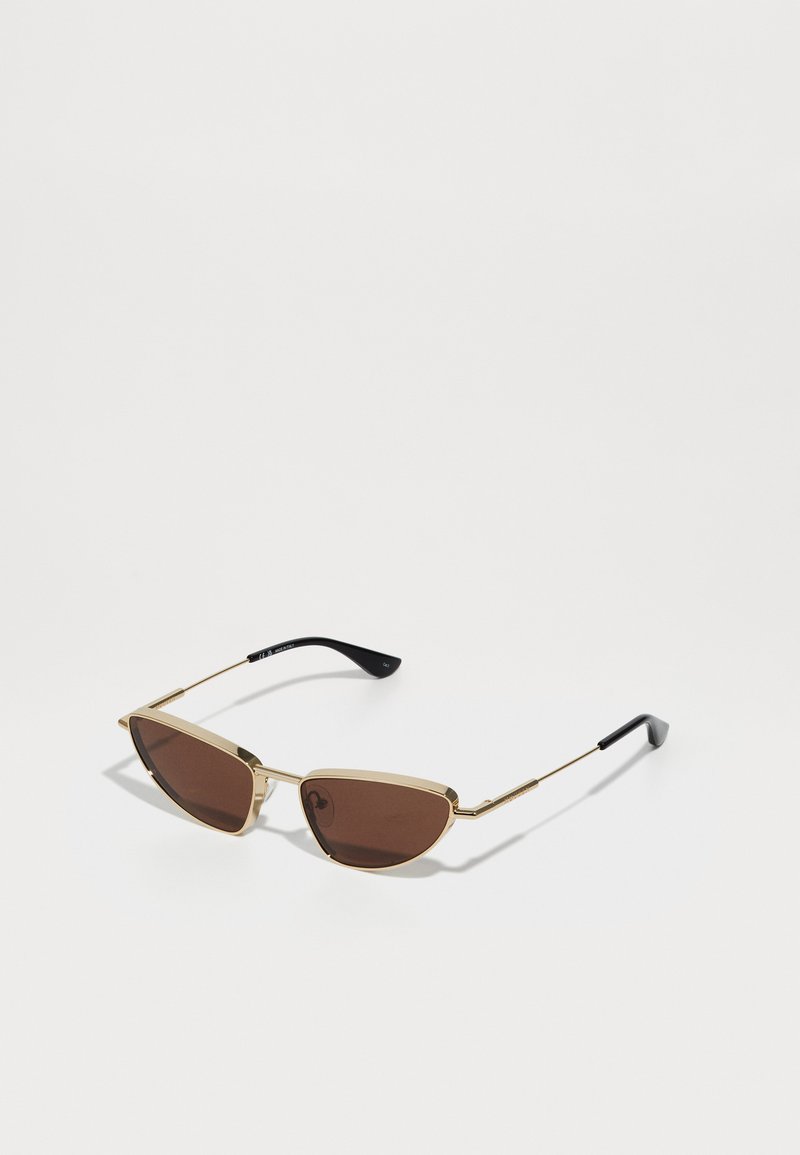 Ochelari de soare cu ramă subțire aurie, lentile maro în formă de ochi de pisică și vârfuri de brad negre, așezati pe o suprafață albă.