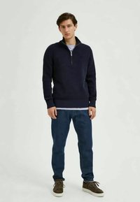 Navy blue zip-up sweater with a high collar, paired with dark jeans and brown lace-up shoes. Textured knit material and relaxed fit.
