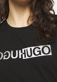Musta puuvillapaita, jossa on hopeinen logo, jossa lukee "HUGO" ja joka on näkyvästi esillä rinnassa. Tasainen pinta, pyöreä kaula-aukko.