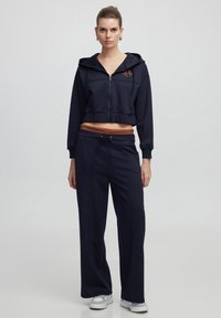 Sudadera corta de azul marino con cremallera y cordón, combinada con pantalones de pierna ancha azul marino con una cinturilla marrón. Textura de tela suave.
