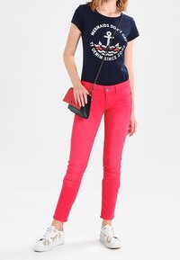 Mujer vistiendo una camiseta gráfica azul marino con ancla, pantalones ajustados rosa brillante, zapatillas blancas con estampado de perros y un pequeño bolso cruzado negro y rojo.
