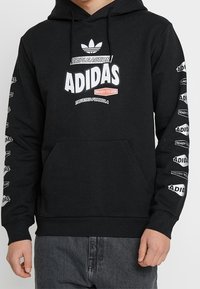 Černá mikina Adidas s bílým a červeným textem "Triple Action," "Adidas," "Ready to Use" a "Trusted Formula" na hrudníku a rukávech.