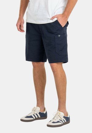Hombre vistiendo pantalones cortos cargo azul marino y zapatillas blancas con rayas azul marino, de pie con una mano en el bolsillo sobre un fondo blanco.