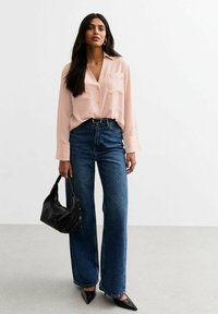 Lichtroze overhemd met knopen en een borstzak, gedragen met hoge taille blauwe wijde jeans. Een zwarte handtas en spitse hakken maken de outfit compleet.