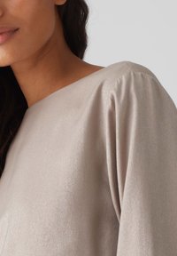 Hellebeige Bluse aus einem glatten, leicht strukturierten Stoff; mit weitem Halsausschnitt und gerüschten Ärmeln. Weicher Glanz und lockere Passform sind zu beobachten.