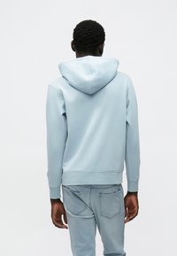 Jasnoniebieska bluza z kapturem, przestronnym kapturem, ściągaczami na mankietach i na dole oraz miękką fakturą. Nosi się ją na jasnoniebieskie spodnie dżinsowe.