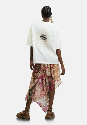 Nő fonott hajjal, fehér, túlméretezett felsőt visel, amelynek hátán nap motívum található, virágmintás aszimmetrikus szoknyát, nagy virág alakú fülbevalót és barna szandált.