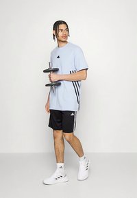 Šviesiai mėlynas marškinėlių su juodais akcentais, juodi šortai su baltomis dryžuotomis detalėmis ir balti sportiniai bateliai, laiko hantelį.