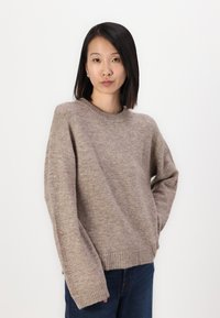 Beige stickad tröja med rund halsringning, avslappnad passform, ribbad mudd och nederkant, mjukt material, bärs med mörkblå jeans.