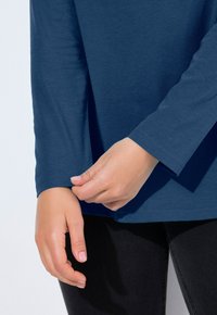 Ręce dostosowujące mankiet długiego rękawa granatowej koszuli, z ciemnymi spodniami widocznymi na jasnym tle.