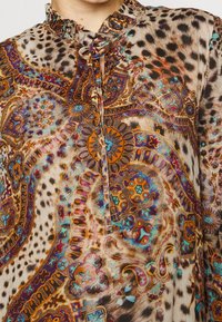 Un gros plan sur le chemisier d'une femme présentant un motif vibrant et complexe avec des designs en cachemire et des motifs animaliers dans des couleurs chaudes.