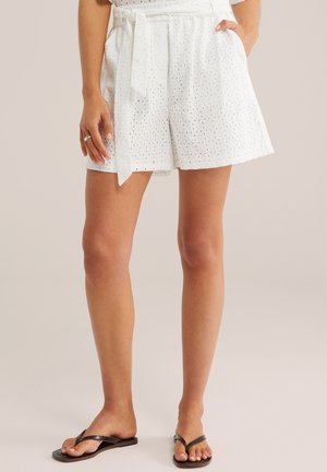 Žena oblečená v bielych šortkách z dierkovaného plátna s viazaným opaskom, stojaca s jednou rukou v vrecku a na nohách čierne žabky.