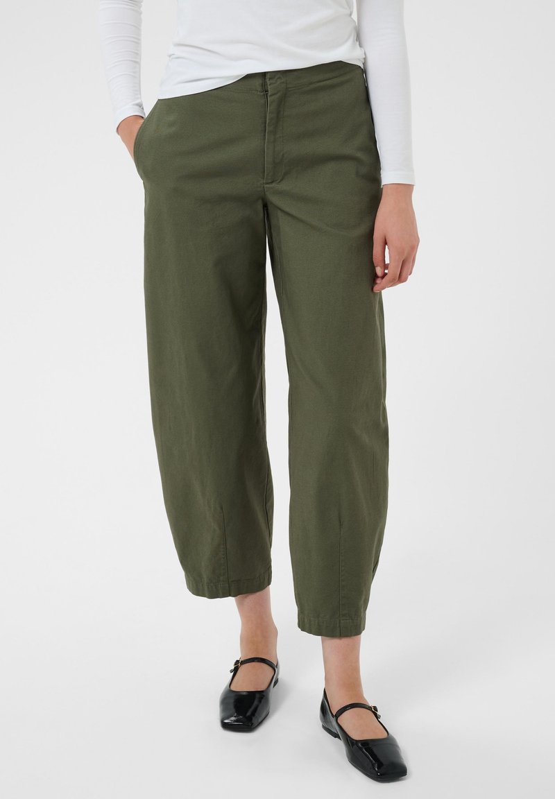 Personne portant un pantalon large court vert olive, un haut blanc à manches longues et des chaussures Mary Jane noires avec une bride et un talon bas.