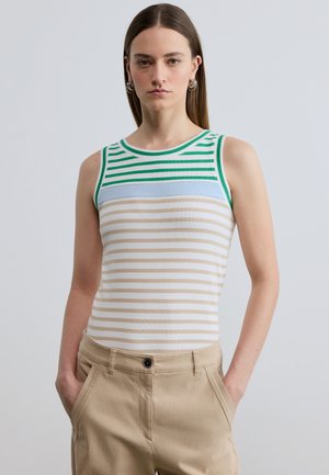 Young woman with straight brown hair wears sleeveless striped top with green, blue, and beige lines, beige pants, and silver hoop earrings, hands in pockets.