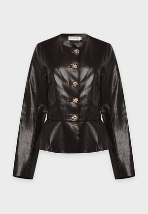 Un mannequin se tient devant un fond clair, vêtu d’une veste sombre en cuir à basque avec des boutons dorés, d’un pantalon noir et de chaussures noires.