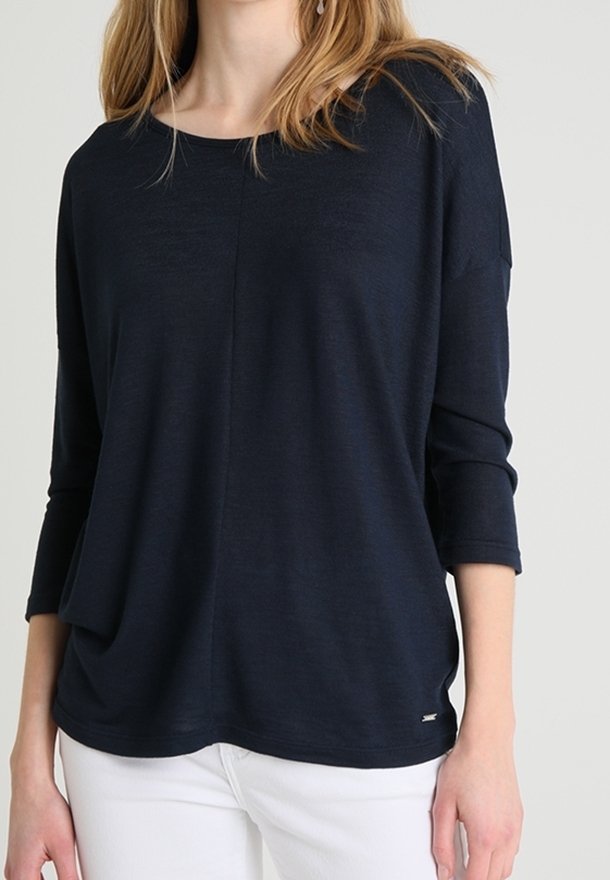 Femme portant un haut noir ample à manches longues avec un col rond et un pantalon blanc, debout devant un fond uni.