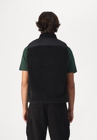 Black fleece vest with a high collar, featuring a textured fabric on the back and smooth material on the shoulders. Worn over a green shirt.