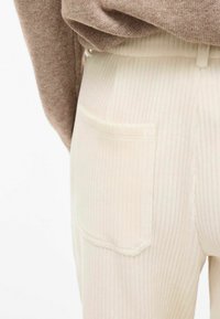 Krämfärgade ribbade corduroy-byxor med en bakficka, med en mjuk textur och subtila vertikala linjer, visade tillsammans med en brun tröja.