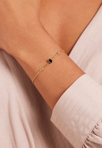 Gouden kettingarmband met een ovale zwarte steen en kleine heldere accenten, gedragen om een pols, met een gestructureerde stofachtergrond.