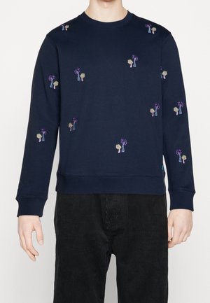 Sweat-shirt à manches longues bleu marine avec petits motifs de champignons brodés en violet et beige, porté avec un pantalon en velours côtelé noir.
