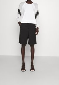 Top blanco con detalles de encaje en las mangas, combinado con pantalones cortos negros hasta la rodilla. La modelo lleva sandalias negras con tiras. Texturas de tela suaves.