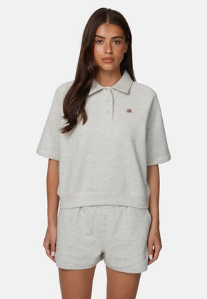 Donna con capelli lunghi e scuri che indossa una felpa grigio chiaro a maniche corte con colletto e pantaloncini coordinati, in piedi davanti a uno sfondo bianco.