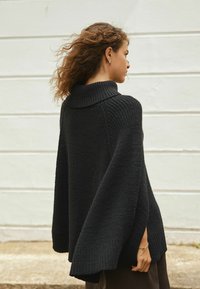 Woman with curly hair wearing a black knitted cape sweater and gold bracelet, standing sideways against a light-colored wall.