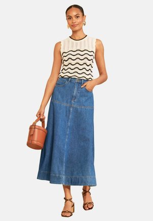 Žena nosi bezrukávový krémový pletený top s čiernym vlnkovým vzorom, modrú denim midi sukňu, hnedé sandále na podpätku a hnedú okrúhlu kabelku.