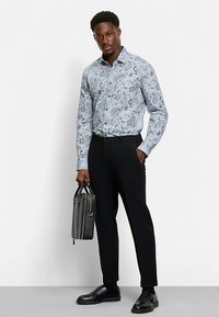 Hellblaues Hemd mit Paisley-Muster, schwarze Hose und schwarze Schuhe, hält eine grau-schwarze Tasche. Einfaches, maßgeschneidertes Design.